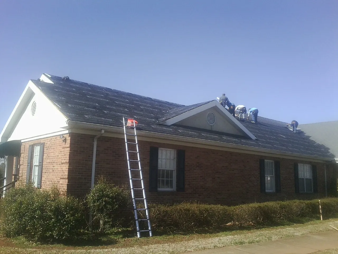 Crew tearing shingles off a brick ranch roof with a ladder set — Metal Roof Repair work by our crew in Deposit